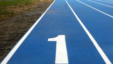 Number 1 / number one lane of running track, white number and white lane lines on blue surface. Low level view with grass runners trees visible in the distance.