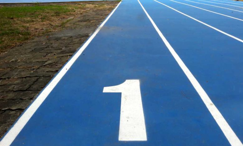 Number 1 / number one lane of running track, white number and white lane lines on blue surface. Low level view with grass runners trees visible in the distance.