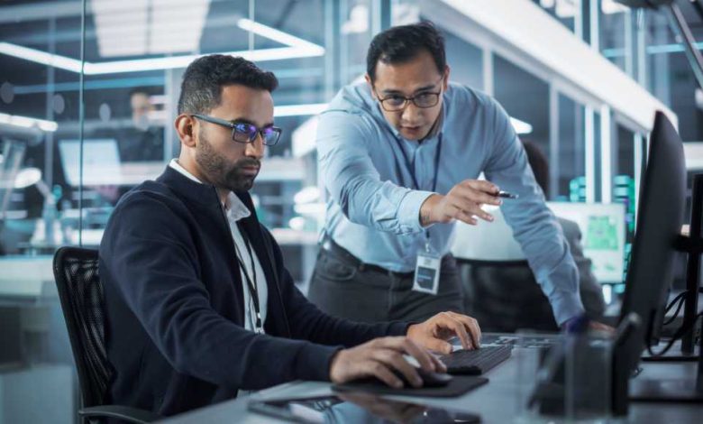 Male Project Supervisor Talks to an Industry 4 Engineer Who Works on Desktop Computer in Robotics Startup Industrial Office. Software Developer Discussing Work Related Tasks During a Meeting.