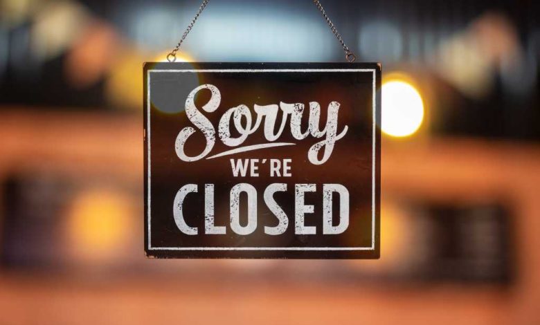 Sorry we're closed sign hanging inside a glass door, colored lights in the background.