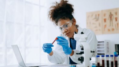 A woman dressed in a white lab coat examines a vial while sitting in front of a computer.